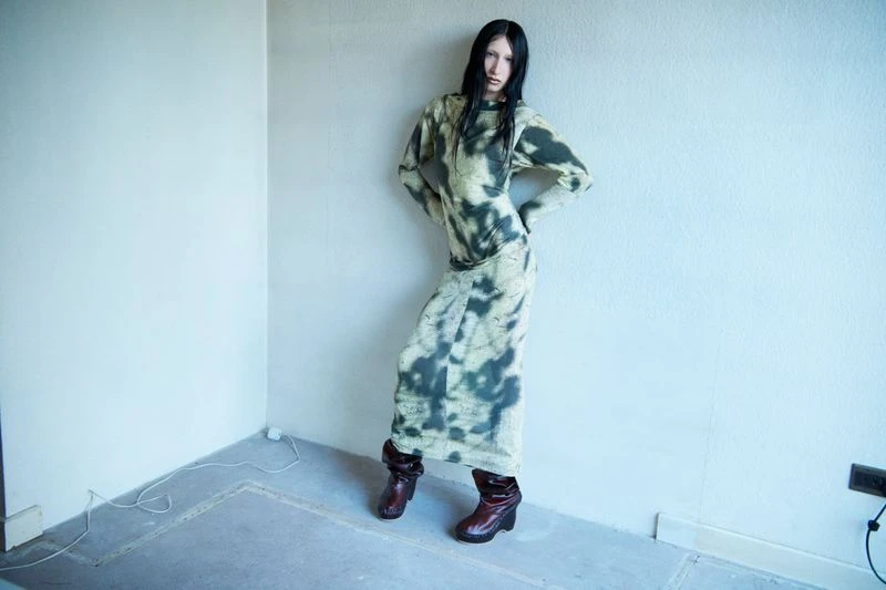 Woman in tie-dye dress and platform boots poses in a corner of an empty room with bare walls and a concrete floor, looking at the camera.