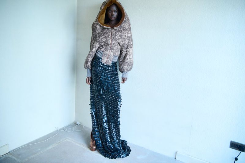 A tall, dark-skinned fashion model poses in an indoor studio, wearing a large, patterned hooded jacket and long, dark, textured skirt.