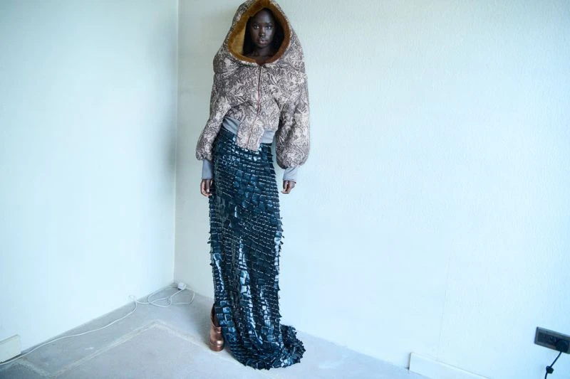 A tall, dark-skinned fashion model poses in an indoor studio, wearing a large, patterned hooded jacket and long, dark, textured skirt. 