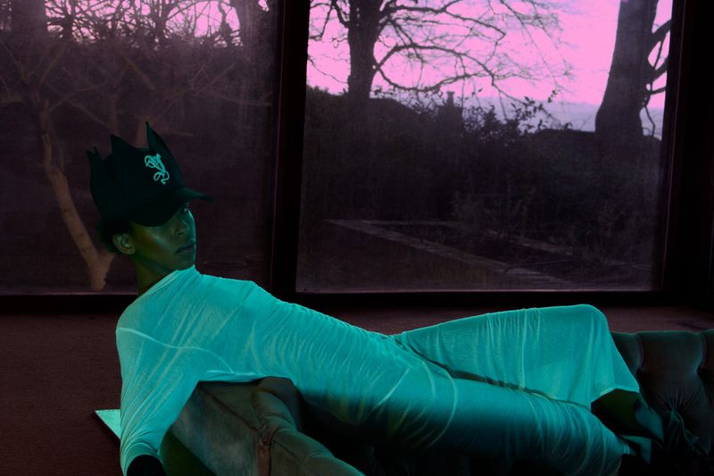 Adult in a surreal indoor fashion portrait, wrapped in a draped cloth, wearing a cap, and lounging on a sofa with outdoor window view.