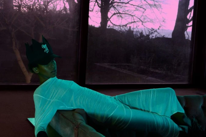 Adult in a surreal indoor fashion portrait, wrapped in a draped cloth, wearing a cap, and lounging on a sofa with outdoor window view.
