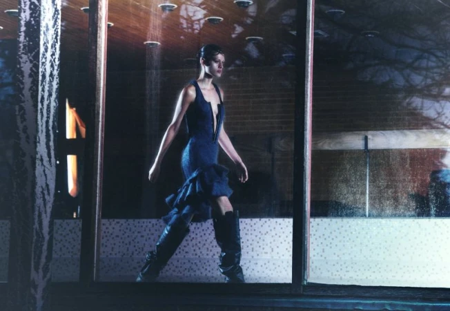 A woman in a blue dress and black boots walks in front of a building with large windows and dark interior, creating a fashion-forward image.