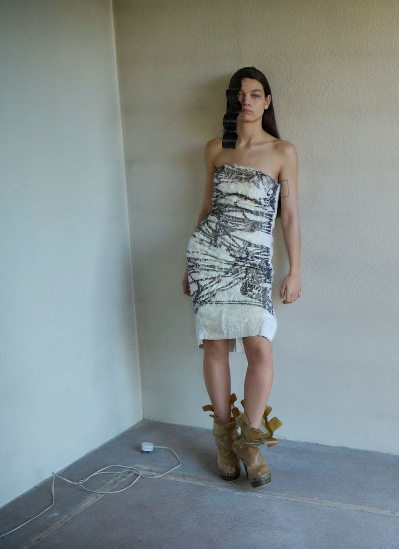 A fair-skinned woman posing indoors against a wall, wearing a strapless dress with platform boots in a fashion shot.