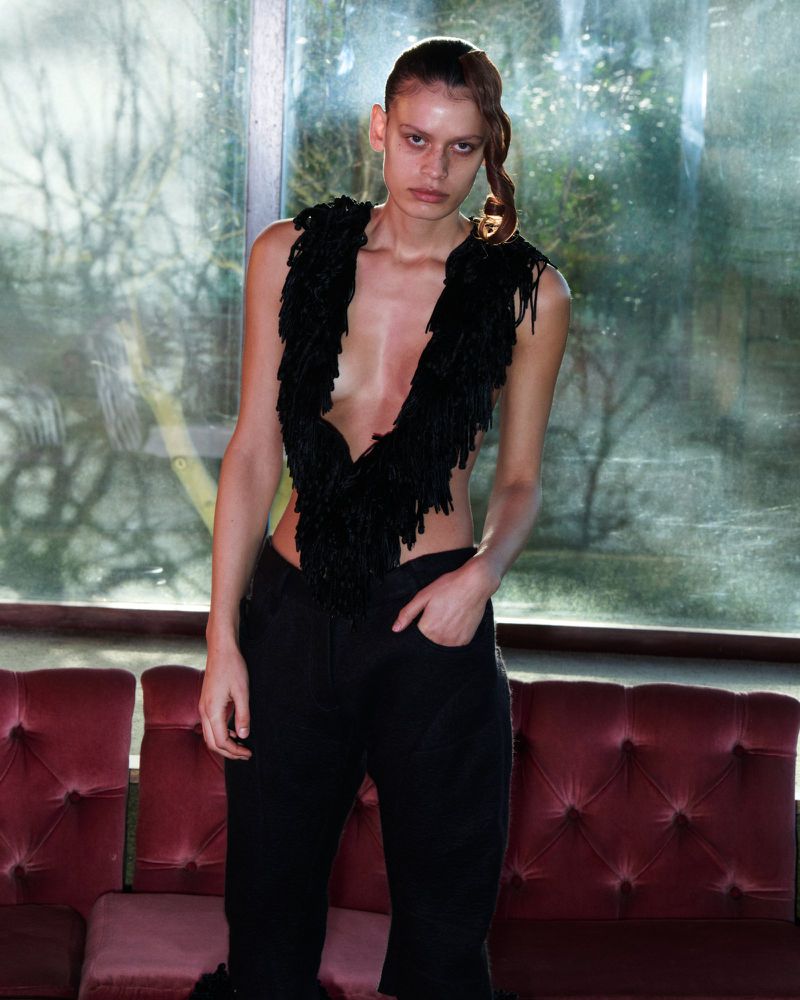 Fashionable young adult woman poses with black fringe top and dark pants against a faded window and velvet couch, medium shot.