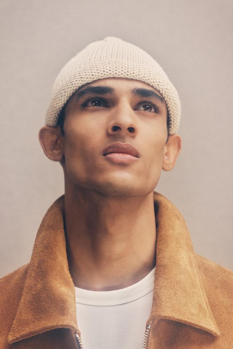 Headshot of a young adult male with dark hair wearing a cream beanie hat and tan jacket looking upwards against a light gray background.