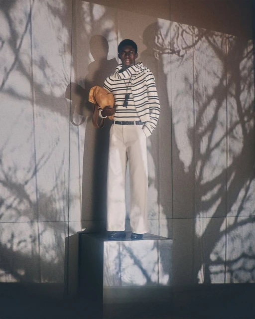 Black male model wearing a striped sweater and white pants stands on a reflective cube in a photo studio with shadows on the wall.
