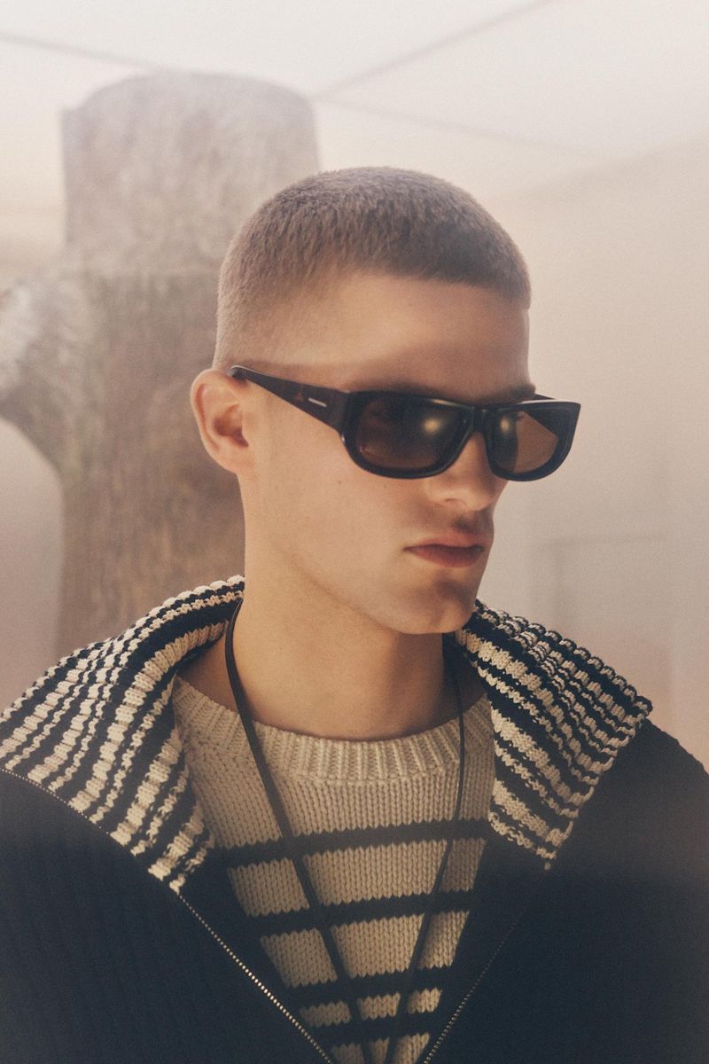A young adult male with a buzzcut wears sunglasses and a striped sweater with a black zip-up jacket, posing in a bright indoor setting.