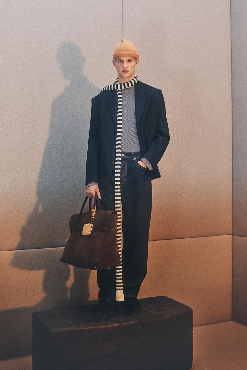 A fair-skinned adult male models a striped scarf, knit hat, black blazer, dark denim pants, and leather satchel on a black block.