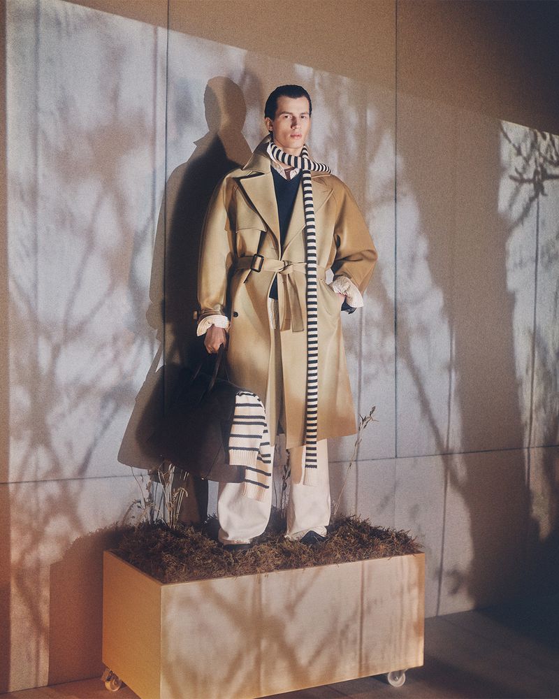 A stylish adult male model stands on a brown box on wheels, holding a dark bag, while wearing a tan trench coat and scarf.