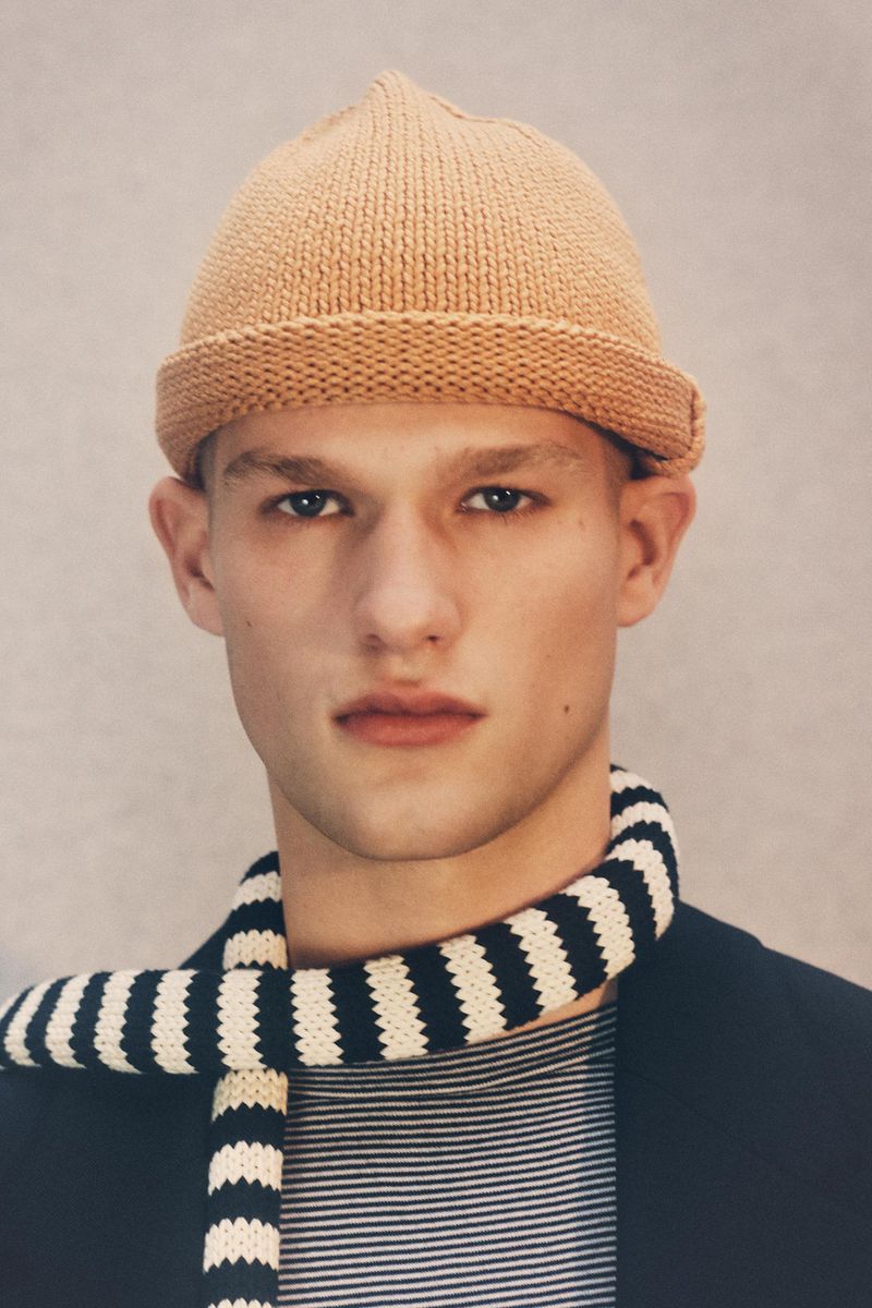 Portrait of a fair-skinned young adult male with a knitted cap and striped scarf, wearing a dark blazer in a neutral indoor setting.