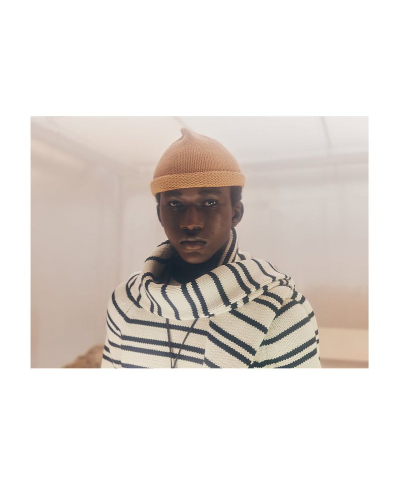 Close-up shot of a stylish African American man indoors wearing a tan beanie and striped knitwear, gazing intently at the camera.
