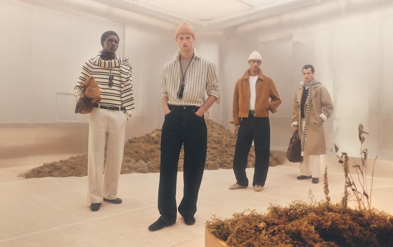 Four adult male fashion models pose indoors in a group wearing casual clothing and accessories in a cool-toned, soft-lit environment.