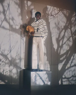 Model standing on a mirrored cube wearing striped sweater and white trousers against a wall with tree branch shadow patterns