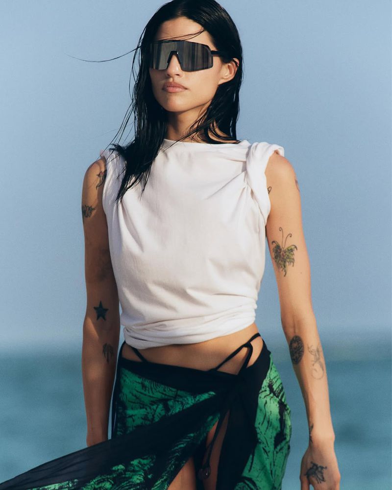 A stylish woman with sunglasses, wearing a white top and sarong, poses on a bright sunny beach, showcasing fashion and lifestyle in a vibrant outdoor setting.