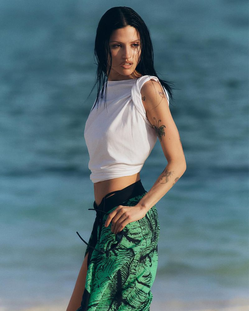 Woman in white crop top and green tropical-print skirt poses on a beach with ocean in background in medium outdoor shot.