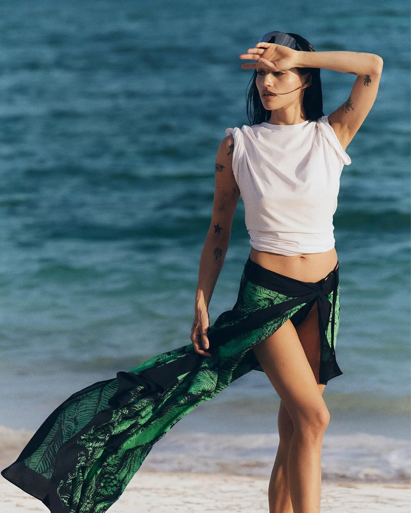 Woman in white shirt and sarong stands on sandy beach with blue ocean in the background, shielding eyes from the sun.