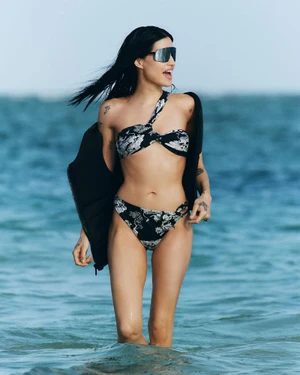 Model wearing a black and white patterned bikini and oversized sunglasses wading in ocean water with a black jacket.