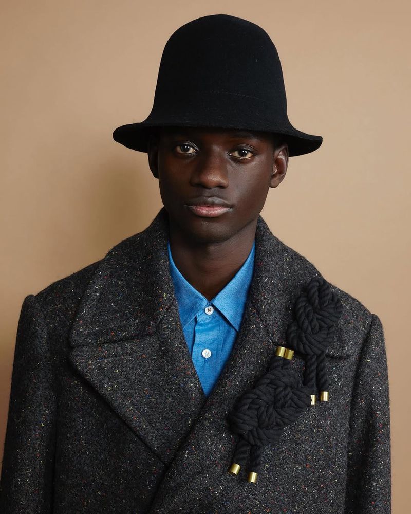 Portrait of a dark-skinned male model wearing a black hat and grey coat with a black rope brooch, posing against a tan background.