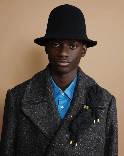 Portrait of a dark-skinned male model wearing a black hat and grey coat with a black rope brooch, posing against a tan background.