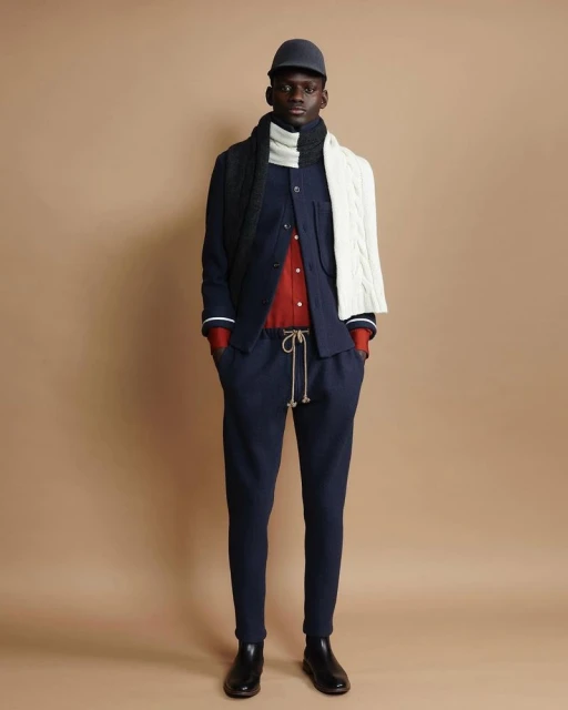 Full-length shot of an adult Black male model in a navy blue suit with a red shirt, a two-tone scarf, a cap, and black boots against a tan background.