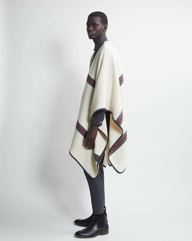 An adult Black male model poses indoors in a gray button-down shirt, gray pants, leather boots, and a cream poncho with gray and red stripes.