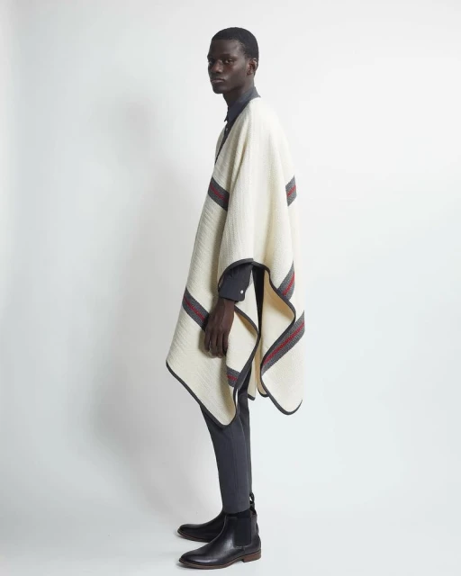 An adult Black male model poses indoors in a gray button-down shirt, gray pants, leather boots, and a cream poncho with gray and red stripes.