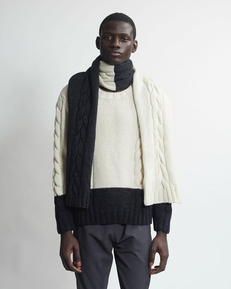 Adult man with neutral expression poses indoors wearing a two-tone cable knit sweater and scarf in ivory and charcoal grey.