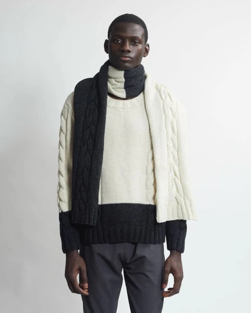 Adult man with neutral expression poses indoors wearing a two-tone cable knit sweater and scarf in ivory and charcoal grey.
