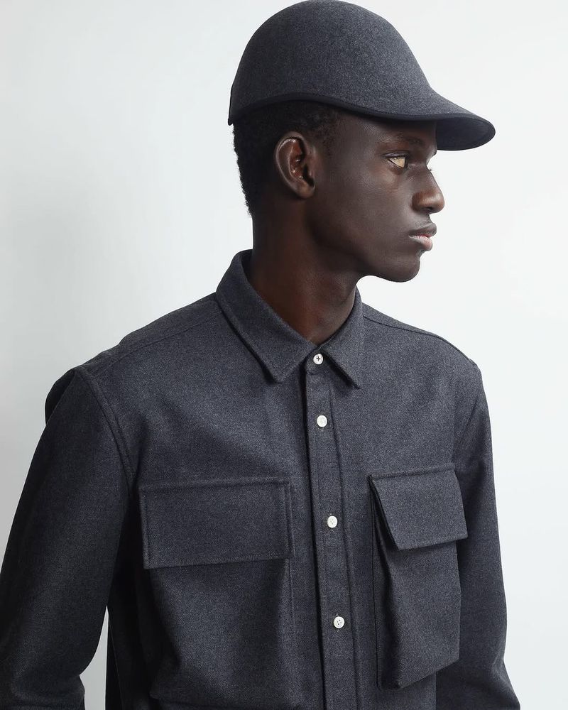 Man in gray shirt and cap poses against white backdrop in a studio shot. Shirt has chest pockets and buttons. Fashion editorial style.