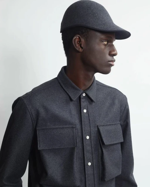 Man in gray shirt and cap poses against white backdrop in a studio shot. Shirt has chest pockets and buttons. Fashion editorial style.