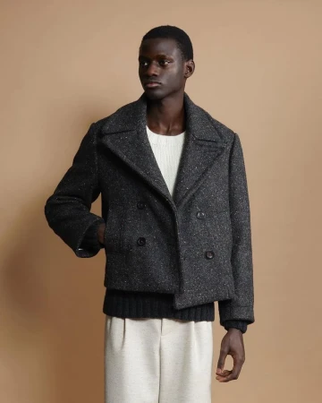 Model wearing a dark speckled wool double-breasted peacoat over a white sweater against a neutral tan backdrop.