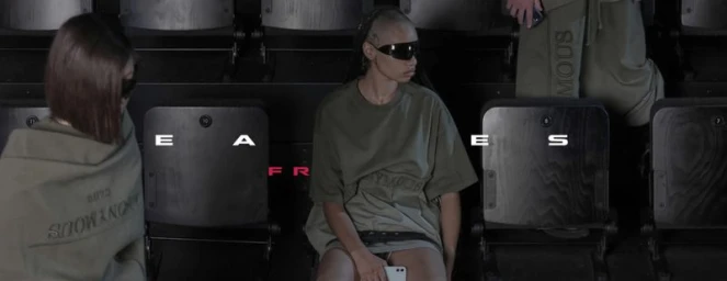 Model wearing dark sunglasses and an olive t-shirt seated in stadium seating for SHAYNEOLIVER collections
