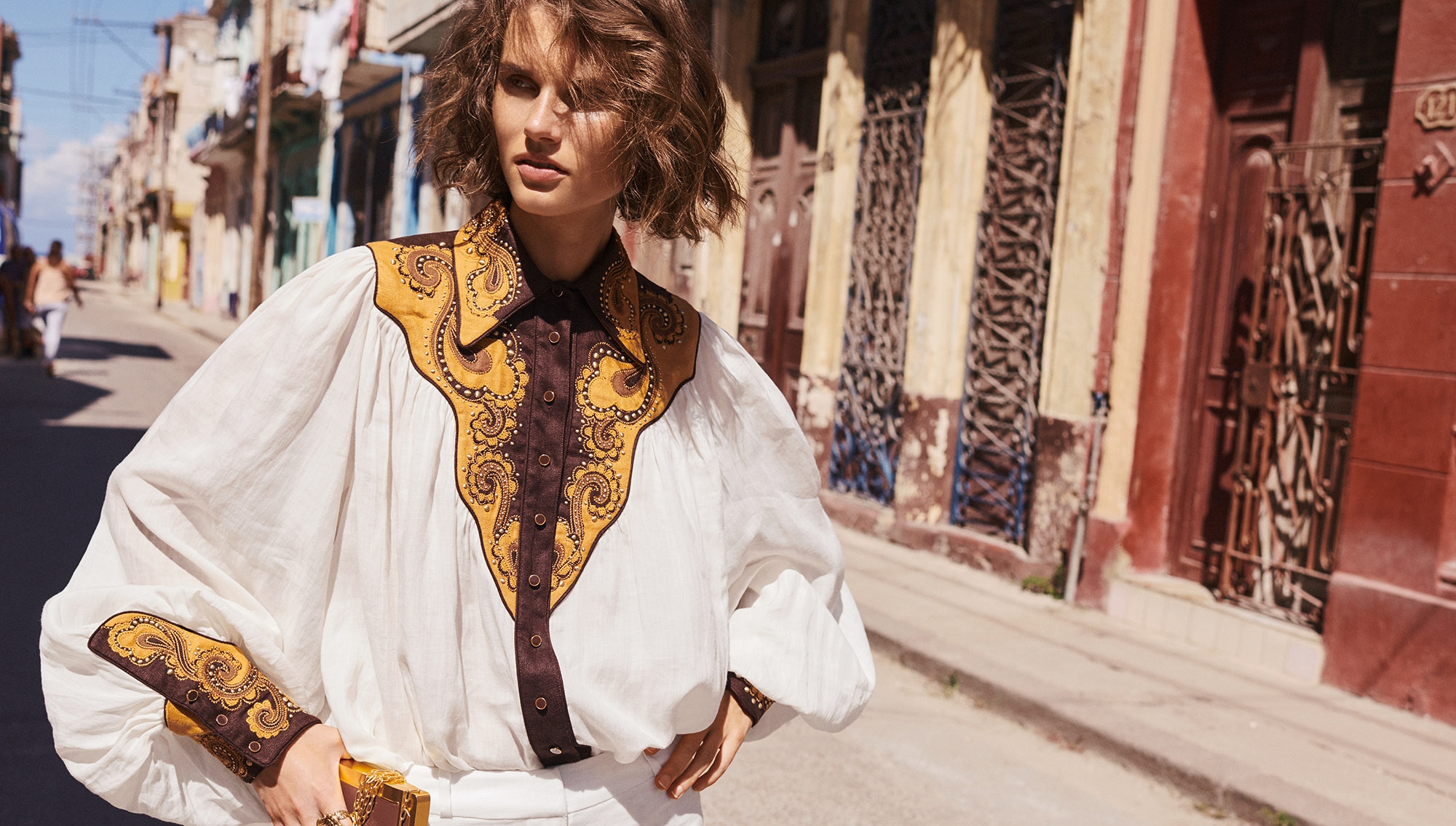 Woman in vintage-style white blouse with brown embroidery, stands on sunlit urban street with old buildings in background.