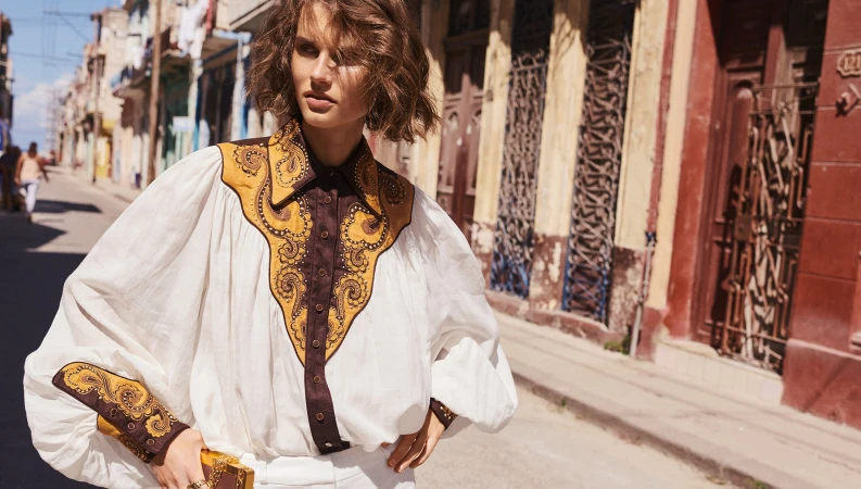Woman in vintage-style white blouse with brown embroidery, stands on sunlit urban street with old buildings in background.