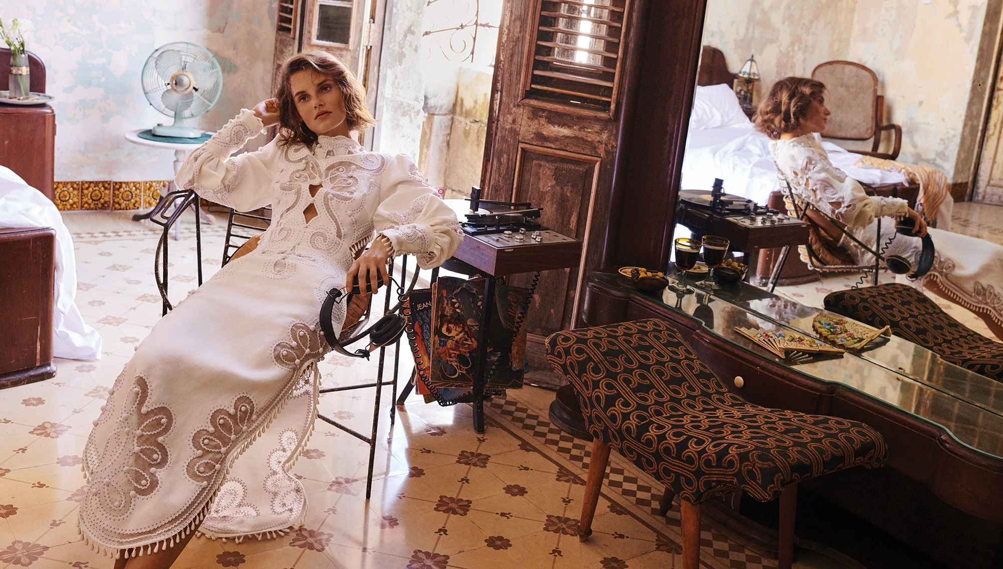 A woman poses in a white lace dress in an indoor setting with vintage decor, captured in a fashion editorial style with soft lighting.