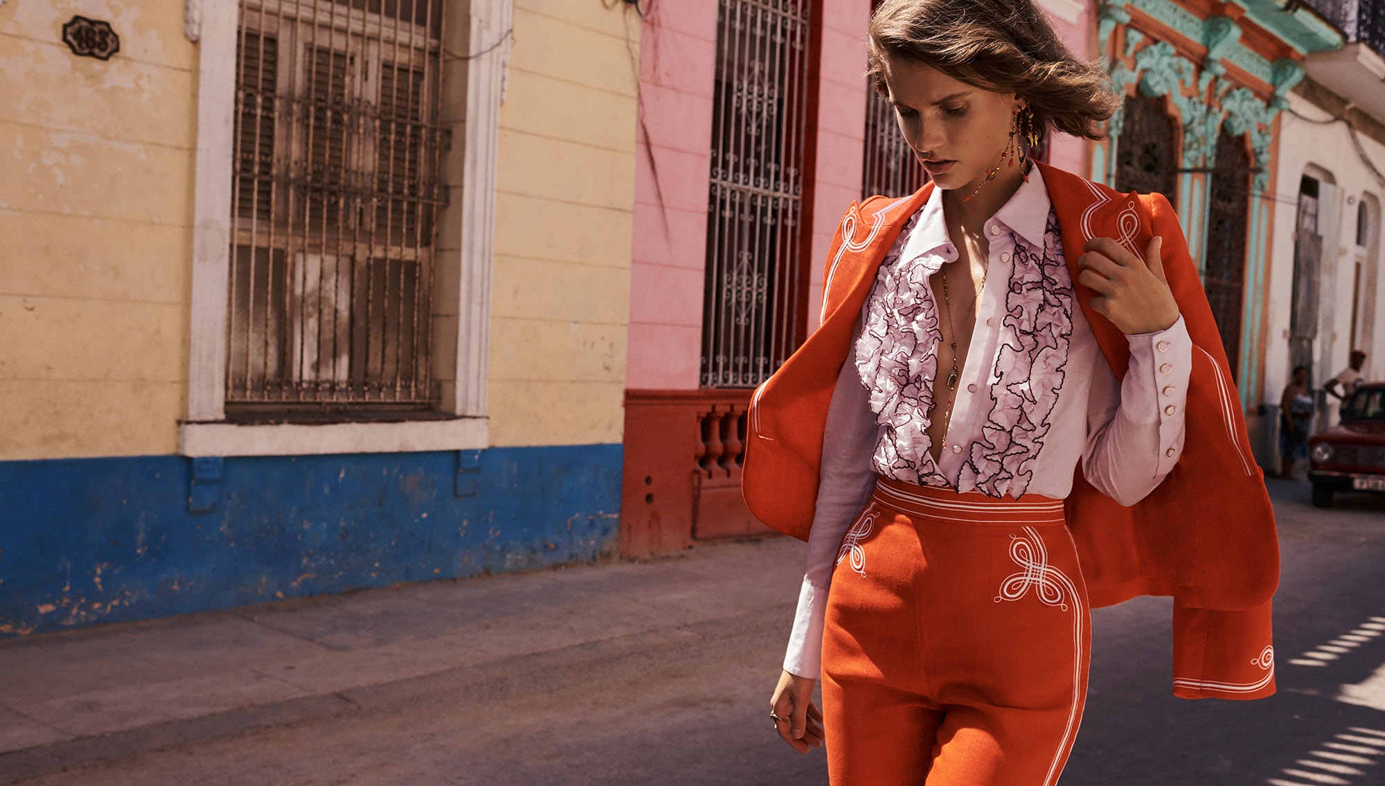 A woman models an orange suit with white detailing in a colorful urban setting, complemented by a ruffled shirt and gold earrings.