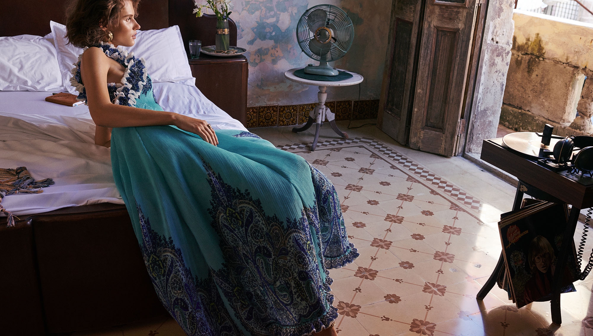 Woman in turquoise dress sits on bed in sunlit room with fan, tiled floor, and vintage decor, near open door with record player.