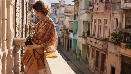 Model in printed silk blouse and tan miniskirt holding a vintage camera on a sunny outdoor balcony overlooking a street