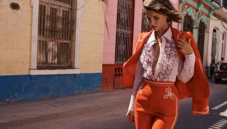 Model in orange suit and ruffled blouse walking past colourful colonial buildings on a sunny street
