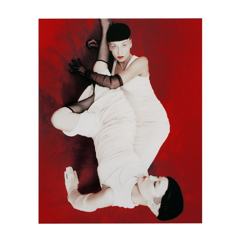 Two women pose in matching white dresses and black hats on a deep red background, captured in a stylish studio shot with dramatic lighting.