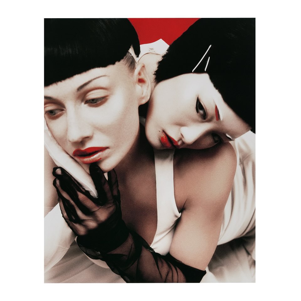 Two women with red lips and short black hair in a close-up portrait, one resting head on the other's shoulder against a red background.