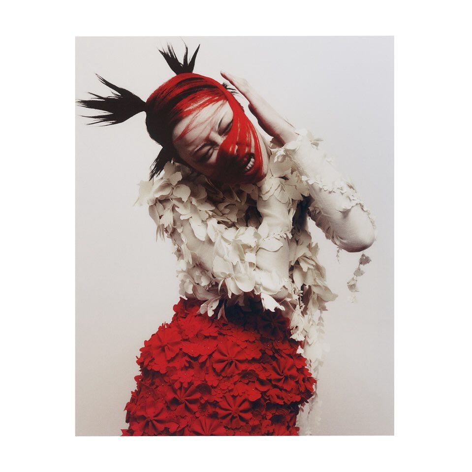 Fashion portrait of a female model with red face paint, spiky hair, white and red layered dress, intense expression, against a light background.