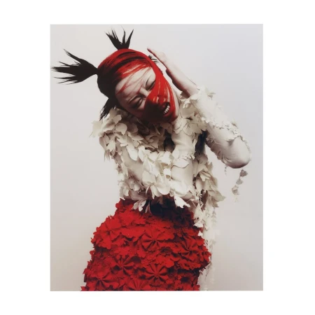 Fashion portrait of a female model with red face paint, spiky hair, white and red layered dress, intense expression, against a light background.