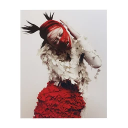 Model with red and black spiky hair and facial paint wearing a textured white top and red skirt against a plain background