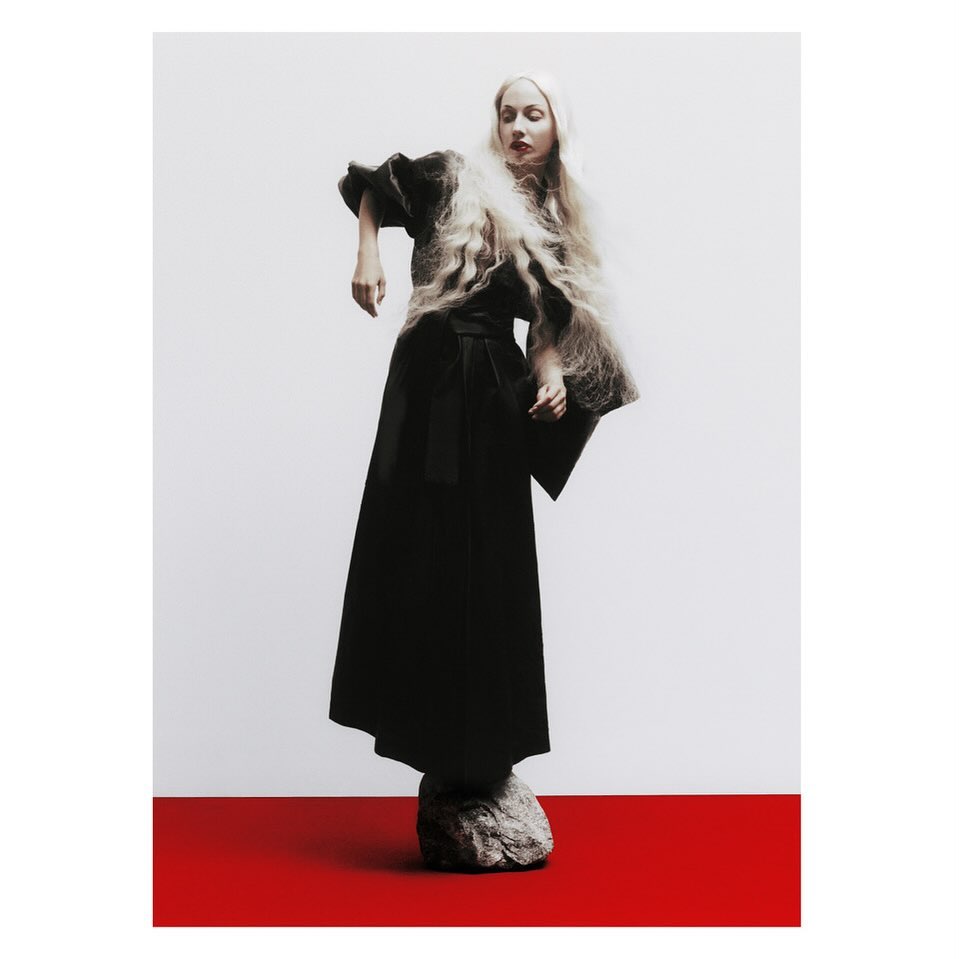 A model in a black gown and voluminous blond hair strikes a dramatic pose atop a grey rock against a red and white backdrop in a studio shot.