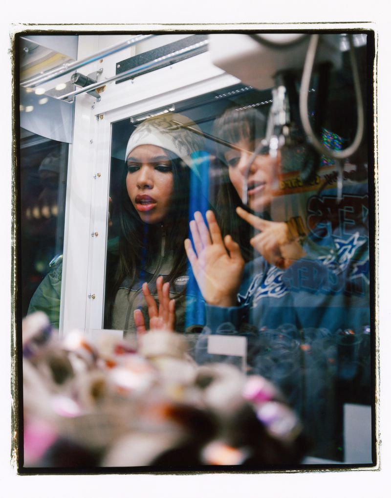Two women peering into a claw machine, looking at the prizes inside, with reflections adding depth to the indoor arcade scene.