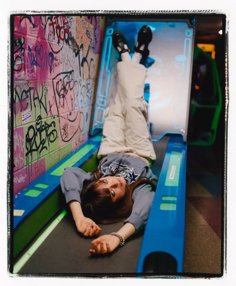 Young woman in casual outfit laying upside down inside an illuminated blue and black arcade game in front of graffiti wall in an indoor setting.