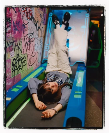 Young woman in casual outfit laying upside down inside an illuminated blue and black arcade game in front of graffiti wall in an indoor setting.
