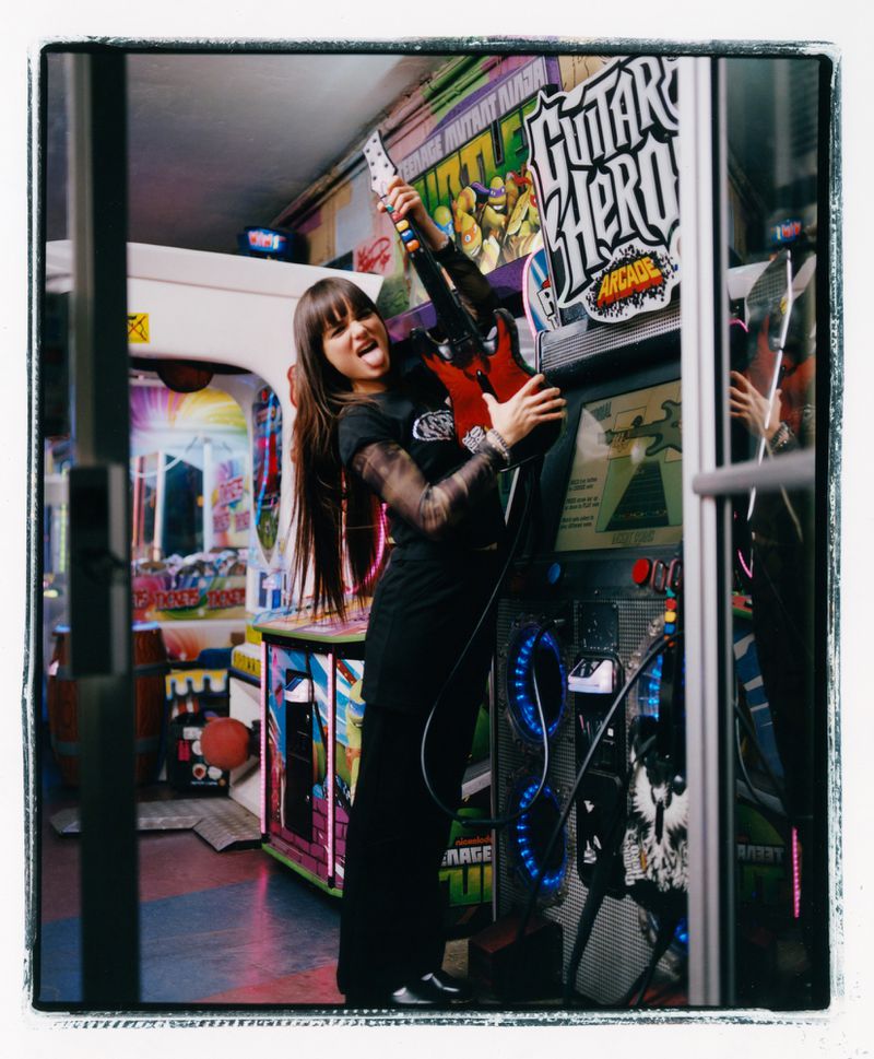 A woman poses with an electric guitar next to a Guitar Hero arcade game in a brightly lit arcade with other games visible.