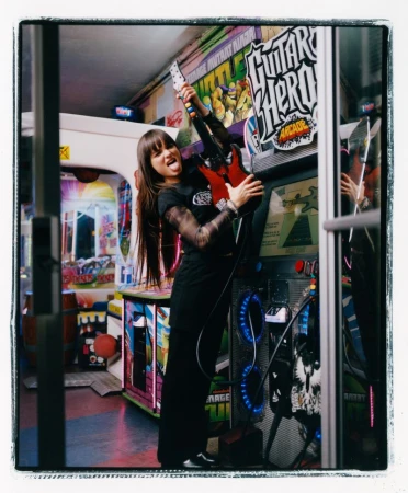 A woman poses with an electric guitar next to a Guitar Hero arcade game in a brightly lit arcade with other games visible.